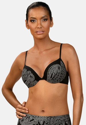 Kobieta ubrana w czarny biustonosz i dopasowaną bieliznę z szarymi wzorami paisley, pozująca z jedną ręką na biodrze, neutralne tło.