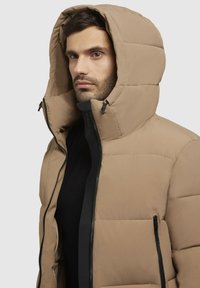 Man met een beige gewatteerde jas met capuchon, zwarte rits en een zwart shirt eronder, tegen een effen grijze achtergrond.