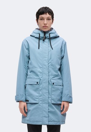 Persona che indossa un cappotto invernale con cappuccio azzurro chiaro, con tasche frontali e cordoncini neri, in piedi davanti a uno sfondo chiaro e semplice.