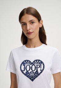 Wit katoenen T-shirt met een navyblauw hartontwerp met florale elementen en de tekst "JOOP!" in het midden. Korte mouwen, ronde hals.