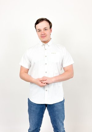 Jeune homme aux cheveux courts et bruns portant une chemise blanche à manches courtes boutonnée et un jean bleu, debout les mains jointes devant lui.