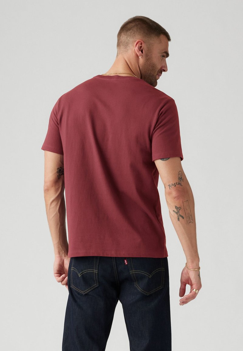 Bordeaux katoenen t-shirt met een ronde hals, korte mouwen en een gladde textuur. Gdragen met donkere denim jeans en zichtbare tatoeages op de arm.