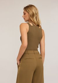 Vrouw met blond haar, gekleed in een mouwloze ribgebreide olijfgroene top en hoge, wijde olijfgroene broek met handen in de zakken, lichtjes zijwaarts gedraaid.