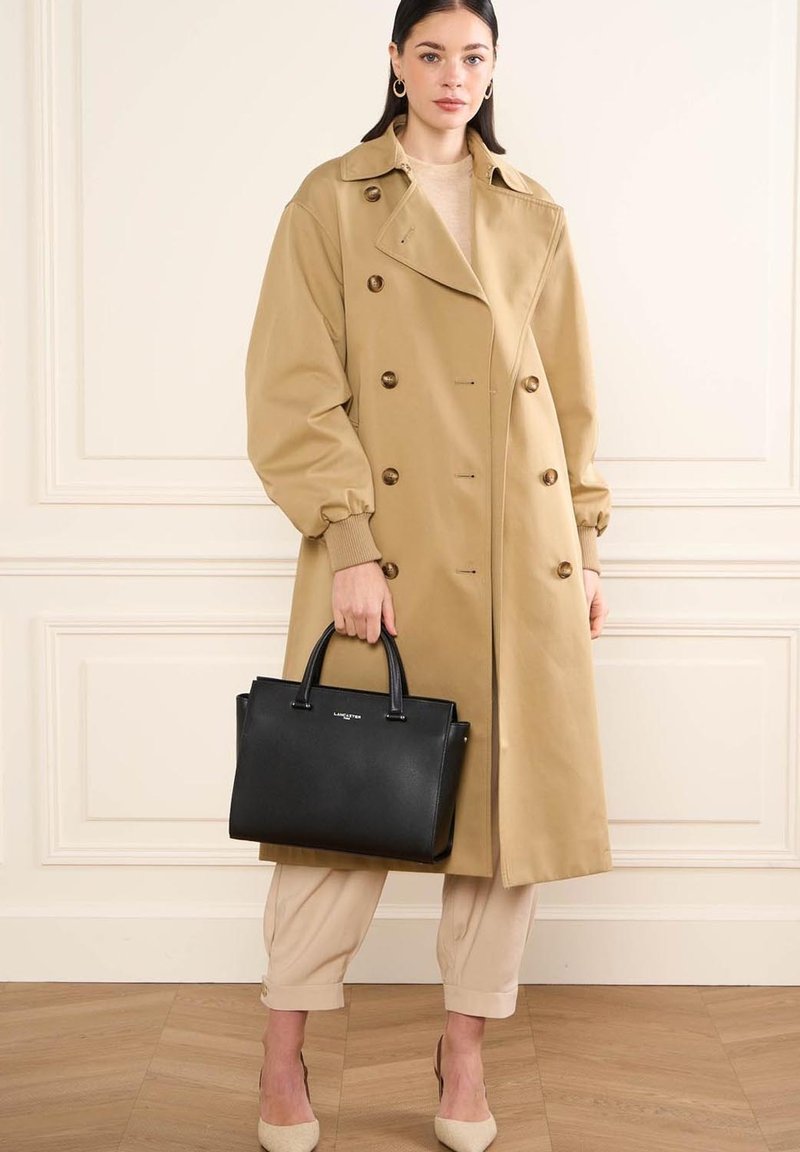 Cappotto trench beige doppiopetto con spalline, borsa nera con manici corti, top crema e pantaloni chiari, tutti su un pavimento di legno.
