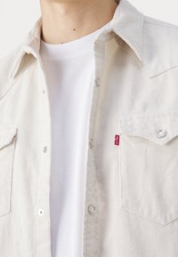 Ljust beige kordskjorta med knappframsida, tryckknappsfickor och silverdetaljer. Bärs över en vit T-shirt med rund halsringning.