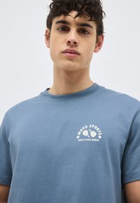 Νεαρός με σγουρά μαλλιά που φοράει μπλε μπλουζάκι με μικρό λευκό αθλητικό λογότυπο στο στήθος, απλό ανοιχτό φόντο.