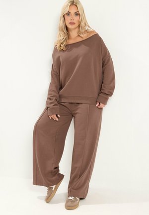 Donna bionda in piedi che indossa un top marrone largo con spalle scoperte, pantaloni larghi coordinati e sneakers beige su uno sfondo chiaro e uniforme.