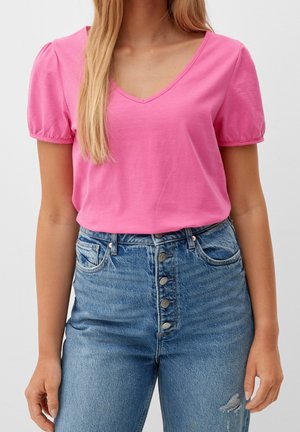 Žena oblečená v růžovém tričku s výstřihem do V a nadýchanými rukávy, zastrčeném do modrých džínů s vysokým pasem a knoflíkovým zapínáním, s mírným opotřebením na stehně.