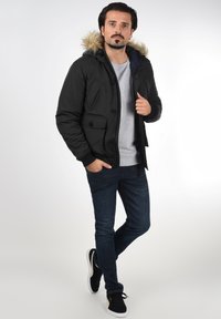 Sort polstret jakke med pelsforet hætte, forknapper og to sidelommer, båret over en grå skjorte og mørke jeans, med sorte sneakers.