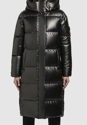 Lange zwarte glanzende gewatteerde pufferjas met capuchon, rits aan de voorkant en zijzakken, gedragen door een staande persoon tegen een effen achtergrond.