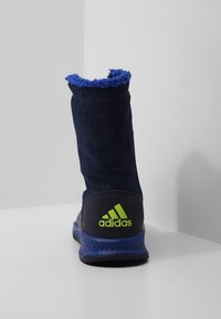 Tmavě modré kotníkové boty s chlupatým modrým límcem, texturovaným svrškem a zeleným logem Adidas na patě. Gumová podrážka s modrými akcenty.