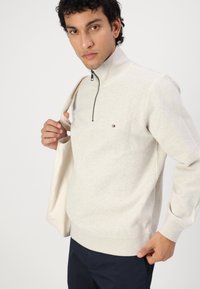 Lichtbeige quarter-zip sweatshirt gemaakt van zachte stof, met een klein logo op de borst en geribbelde manchetten en zoom. Model houdt een tote bag vast.