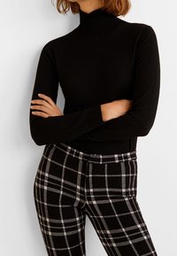 Osoba ubrana w czarny długi sweter z kołnierzem i czarno-białe spódnico-spodnie, z rękami skrzyżowanymi na talii.