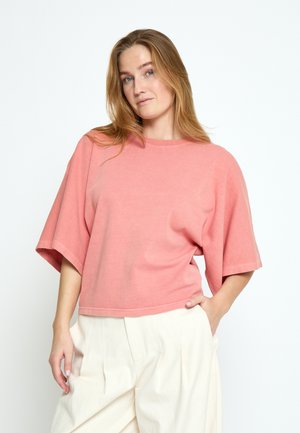 Žena s dlhými blond vlasmi v ľahkom koralovo-ružovom tričku s krátkym rukávom a bielych plisovaných širokých nohaviciach, ruka v vrecku.