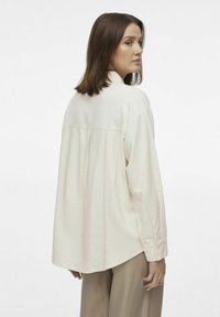 Blusa de manga larga en crema claro con un ajuste holgado, textura suave, detalle de pliegue en la parte trasera y dobladillo redondeado. Lleva puesta con pantalones beige claros.