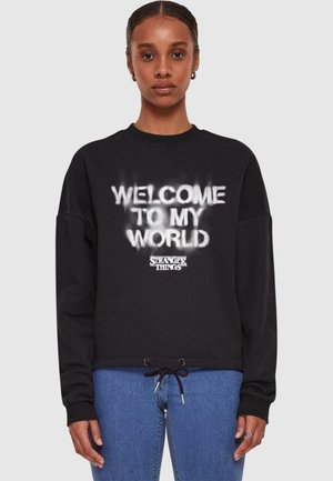 Black sweatshirt featuring "WELCOME TO MY WORLD" in white spray-painted text and "STRANGER THINGS" logo, with a drawstring hem.