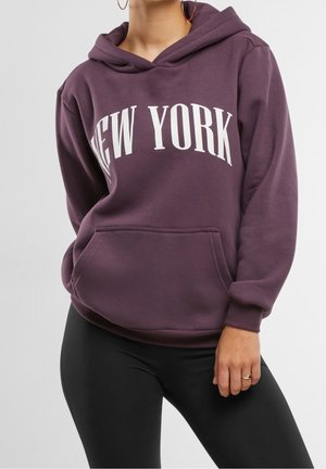 Personne portant un sweat à capuche violet avec "NEW YORK" en lettres blanches et un legging noir, montrant le haut du corps et la main droite avec une bague.