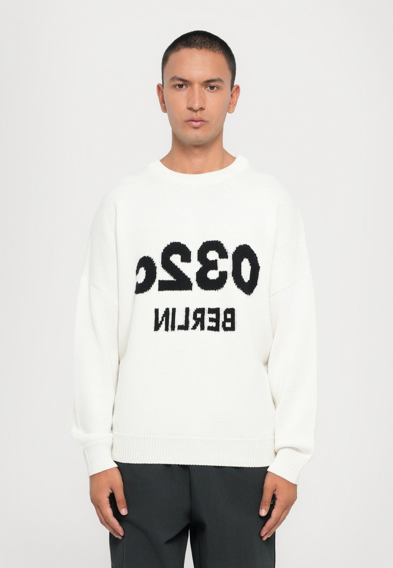 Pull en tricot blanc avec texte noir "0320 BERLIN". Il a un col rond, des manches longues, des poignets et un ourlet côtelés. Texture douce, coupe décontractée.