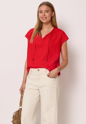 Jeune femme en blouse rouge à manches courtes et pantalon crème à jambes larges tient un sac marron à franges, souriant sur un fond clair.