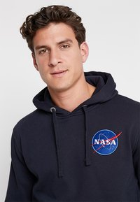 Hanorac cu glugă de culoare albastru marin, realizat din bumbac, având un patch circular cu logo-ul NASA, accentuat cu roșu și un guler cu șnur.