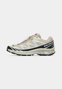 Ανοιχτό μπεζ αθλητικό παπούτσι με επάνω μέρος από δίχτυ, μαύρες λεπτομέρειες, λευκές ρίγες και υφή σόλας. Διαθέτει κλείσιμο με γρήγορο κορδόνι και ενισχυμένη μύτη.