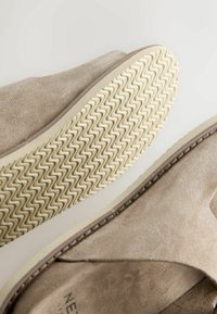 Chaussure en daim beige avec une semelle texturée présentant un motif en zigzag. Le design comprend une couture le long du bord et une forme arrondie.