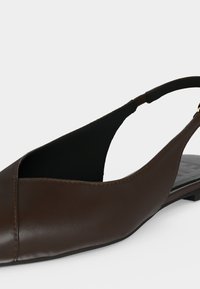 Καφέ δερμάτινο επίπεδο παπούτσι slingback με μυτερό μύτη και μαύρο ελαστικό λουράκι στη φτέρνα σε λευκό φόντο.