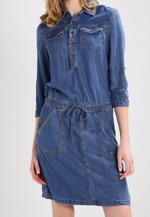 Mujer que lleva un vestido vaquero azul de manga larga hasta la rodilla, con bolsillos en el pecho con botones y cintura con cordón ajustable.