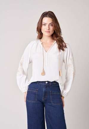 Vrouw met lang bruin haar die een witte geborduurde blouse met franjes en donkerblauwe hooggetailleerde jeans draagt, staand tegen een effen achtergrond.