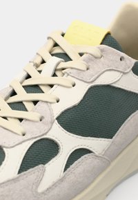 Sneaker med grønt netting overdel, kremfarget lærdetaljer, grå semsket skinn og gul logoetikett på tungen. Har fremtredende snøring og rund form.