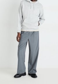 Lichtgrijze hoodie met een kangoeroezak, gecombineerd met wijde grijze broeken en zwarte schoenen. Glad materiaal met minimale naden en neutrale tinten.