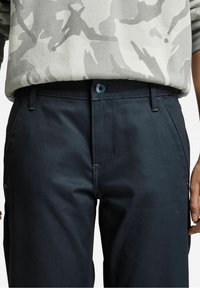 Donkerblauwe marinebroek met een gladde textuur, standaard pasvorm, voorzakken en een blauwe knoopsluiting. Gecombineerd met een lichtgrijs top met camopatroon.