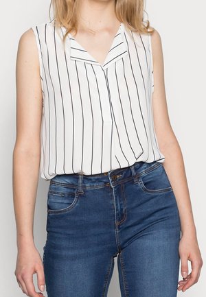 Kvinde iført en ærmeløs hvid bluse med sorte lodrette striber og blå højtaljede jeans står mod en ensfarvet baggrund.