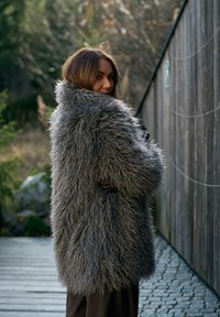 Kobieta z długimi brązowymi włosami nosi oversize szary, puszysty futrzany płaszcz, stojąc na zewnątrz obok drewnianej ściany na kamiennej i drewnianej ścieżce.