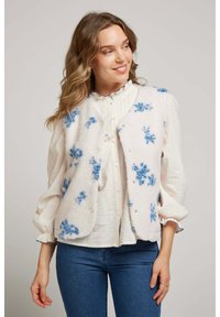 Gilet en polaire blanc avec broderie florale bleue, présentant un col rond, des boutons-pression et une coupe décontractée, superposé sur un haut léger.