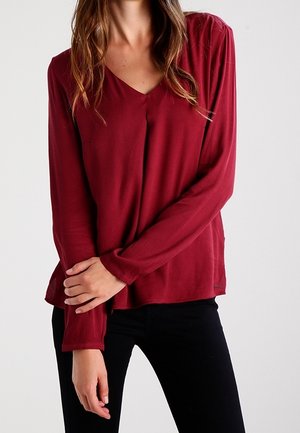 Vrouw die een wijnrode blouse met lange mouwen en V-hals draagt en zwarte broek, staand met één arm gekruist en hand die op de andere arm rust.