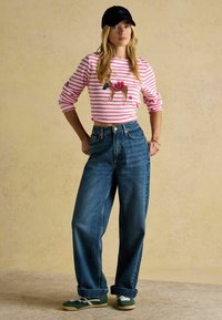 Prúžkované ružové a biele tričko s dlhým rukávom a vyšívanou grafikou, vysokozúne modré džíny a zelené tenisky s bielymi akcentmi.