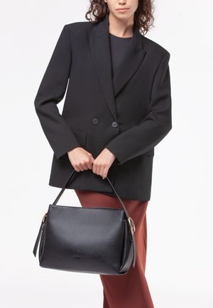 Borsa a mano in pelle nera con un design strutturato, texture liscia e dettagli hardware in tonalità oro, tenuta da una persona in un blazer nero.