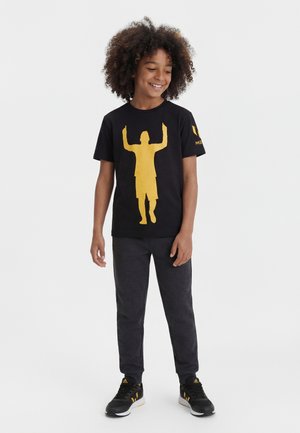 Smilende barn med krøllet hår iført sort t-shirt med gul silhuet, mørke bukser og sorte sneakers mod ensfarvet baggrund.