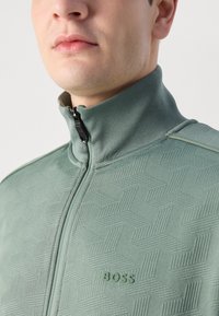 Grønn zip-up jakke med teksturmønster, høy krage og merkevareaccent. Materialet ser glatt ut med en strukturert design.