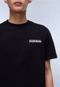 Juodas medvilnės marškinėliai su apvaliu iškirpimu, kuriuose ant kairiojo krūtinės krašto yra balta prekės ženklo logo. Trumpomis rankovėmis, lygaus paviršiaus.