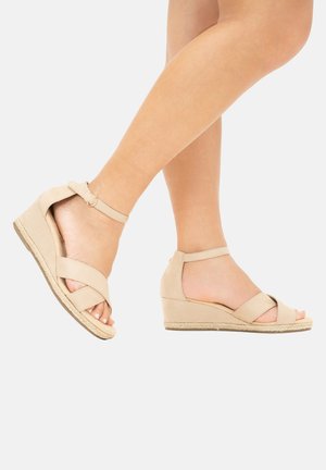 Beige wedge sandals with a suede-like texture, featuring cross-strap design, ankle strap with buckle, and a woven jute accent on the sole.