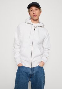 Helle graue Zip-Hoodie mit Kordeln, ausgestattet mit einem Frontreißverschluss und Kängurutaschen. Kombiniert mit locker sitzenden blauen Jeans.