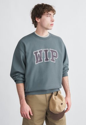 Sudadera gris con "WIP" impreso en púrpura oscuro, puños acanalados y cuello redondo. Gorra beige colgando del cinturón de pantalones marrones.