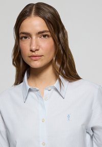 Ljusblå randig knapp-up skjorta i mjukt tyg. Har en liten broderad logotyp på vänster bröst och standardkrage.