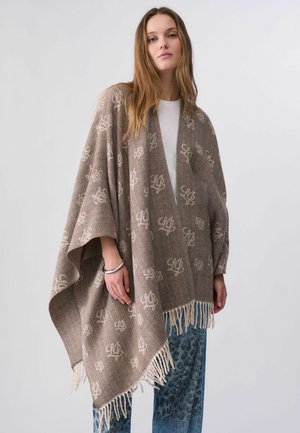 Jonge vrouw met lang haar die een bruin poncho met patroon en franjes draagt over een wit topje en blauwe broek tegen een effen achtergrond.