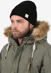 Olijfgroene parka met nepbont gevoerde capuchon, voorzien van beige aantrekkoorden en een ritszak, gecombineerd met een zwarte geribde beanie.