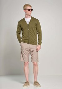 Cardigan verde oliva con bottoni, t-shirt bianca, pantaloni corti beige e scarpe marrone chiaro; modello indossa anche occhiali da sole scuri.