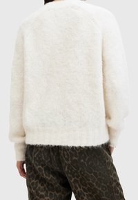 Weißer, flauschiger Pullover mit einer strukturierten Oberfläche, der einen lockeren Schnitt, einen gerippten Saum und Raglanärmel aufweist, kombiniert mit dunkel gemusterten Hosen.