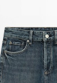 Donkerblauwe spijkerbroek taille met voorzakken, riemlussen, knoopsluiting en stikdetails op een witte achtergrond.